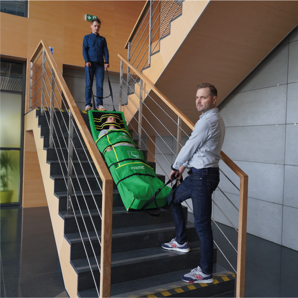 Dwóch mężczyzn stoi na schodach i trzyma pasy od materaca ewakuacyjnego Prime. Na schodach leży mężczyzna zapięty w tym materacu.